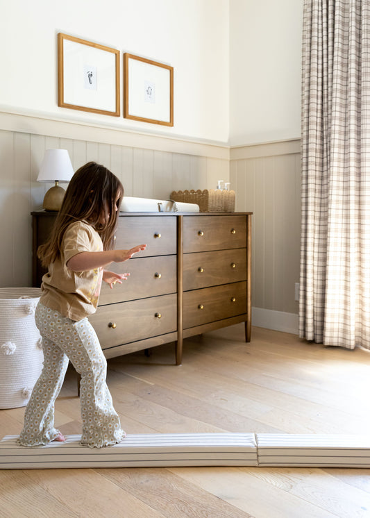 Child practicing balance on a North&Nova beam in a bright bedroom with light wood floors, a dresser, and tall patterned curtains.